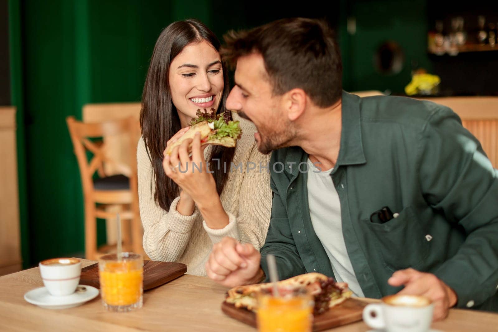 Couple shares laughter and delicious food while sitting at a wooden table in a bright cafe, surrounded by greenery and other patrons enjoying their meals