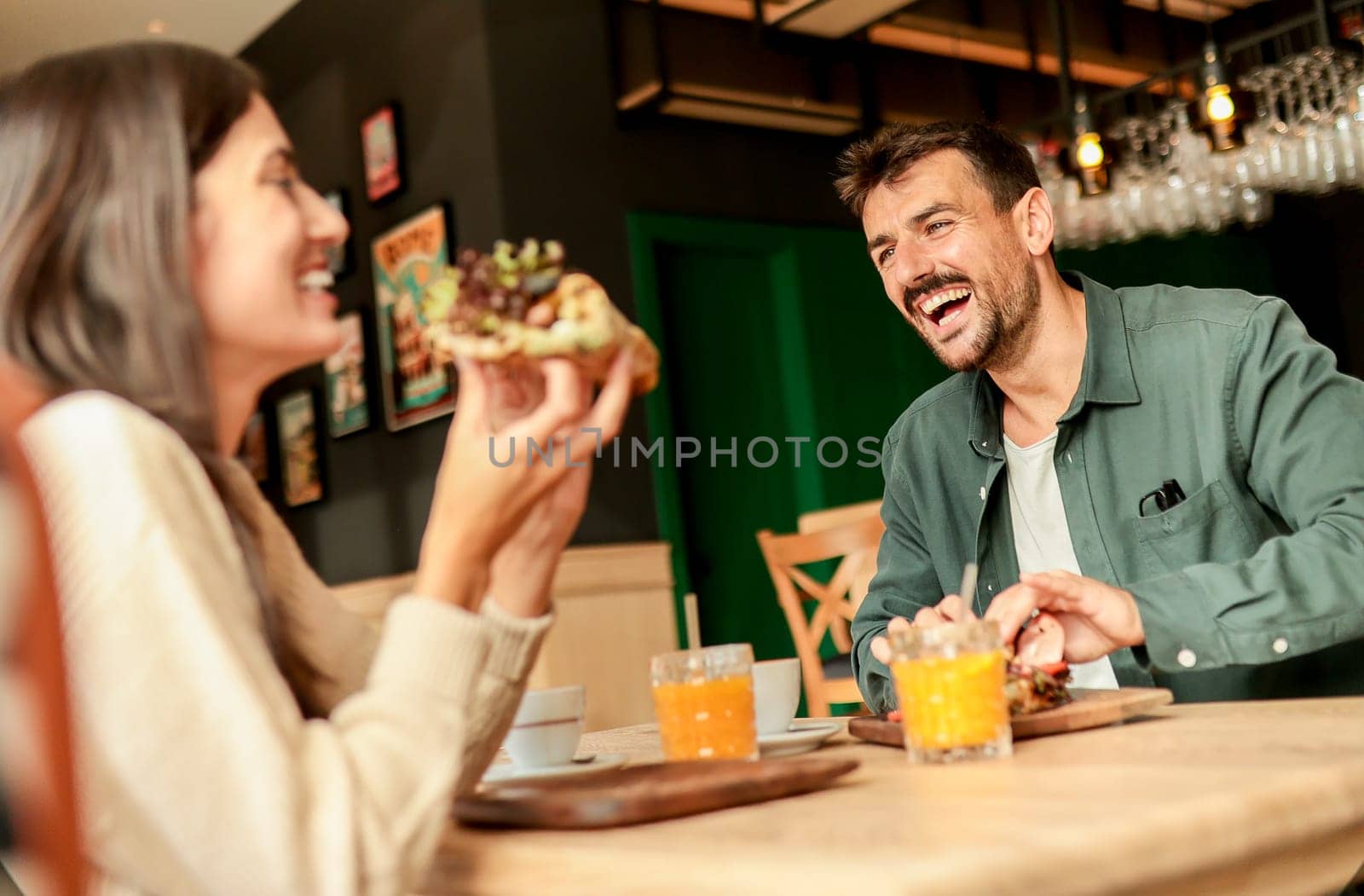 Couple shares laughter and delicious food while sitting at a wooden table in a bright cafe, surrounded by greenery and other patrons enjoying their meals