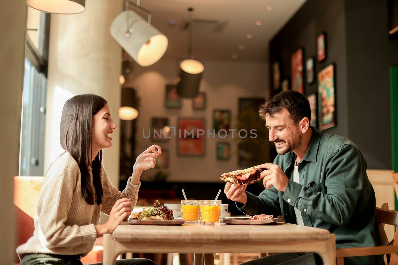 Couple shares laughter and delicious food while sitting at a wooden table in a bright cafe, surrounded by greenery and other patrons enjoying their meals