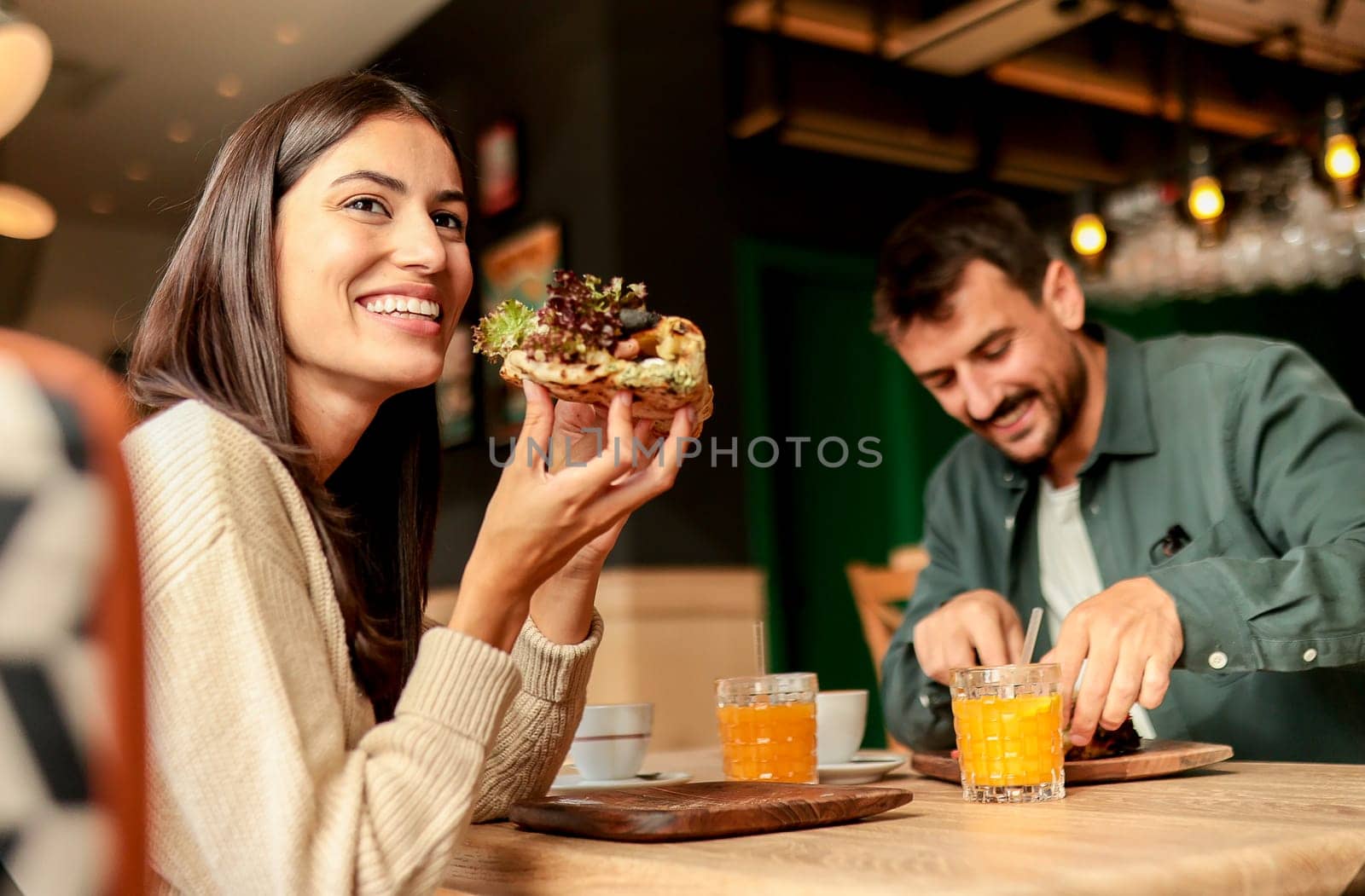 Couple shares laughter and delicious food while sitting at a wooden table in a bright cafe, surrounded by greenery and other patrons enjoying their meals