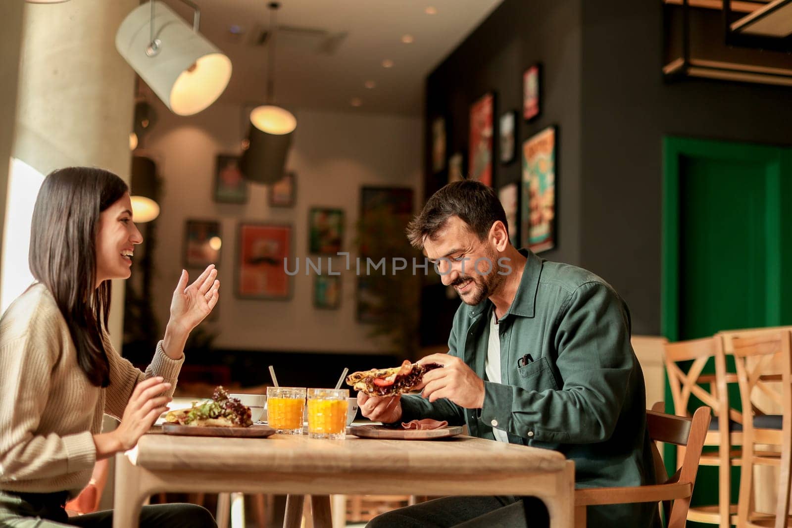 Couple shares laughter and delicious food while sitting at a wooden table in a bright cafe, surrounded by greenery and other patrons enjoying their meals