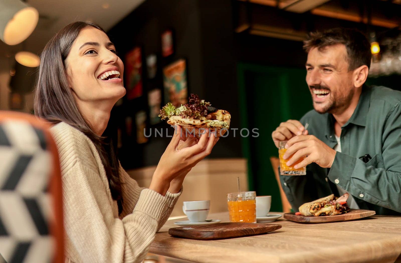 Couple shares laughter and delicious food while sitting at a wooden table in a bright cafe, surrounded by greenery and other patrons enjoying their meals