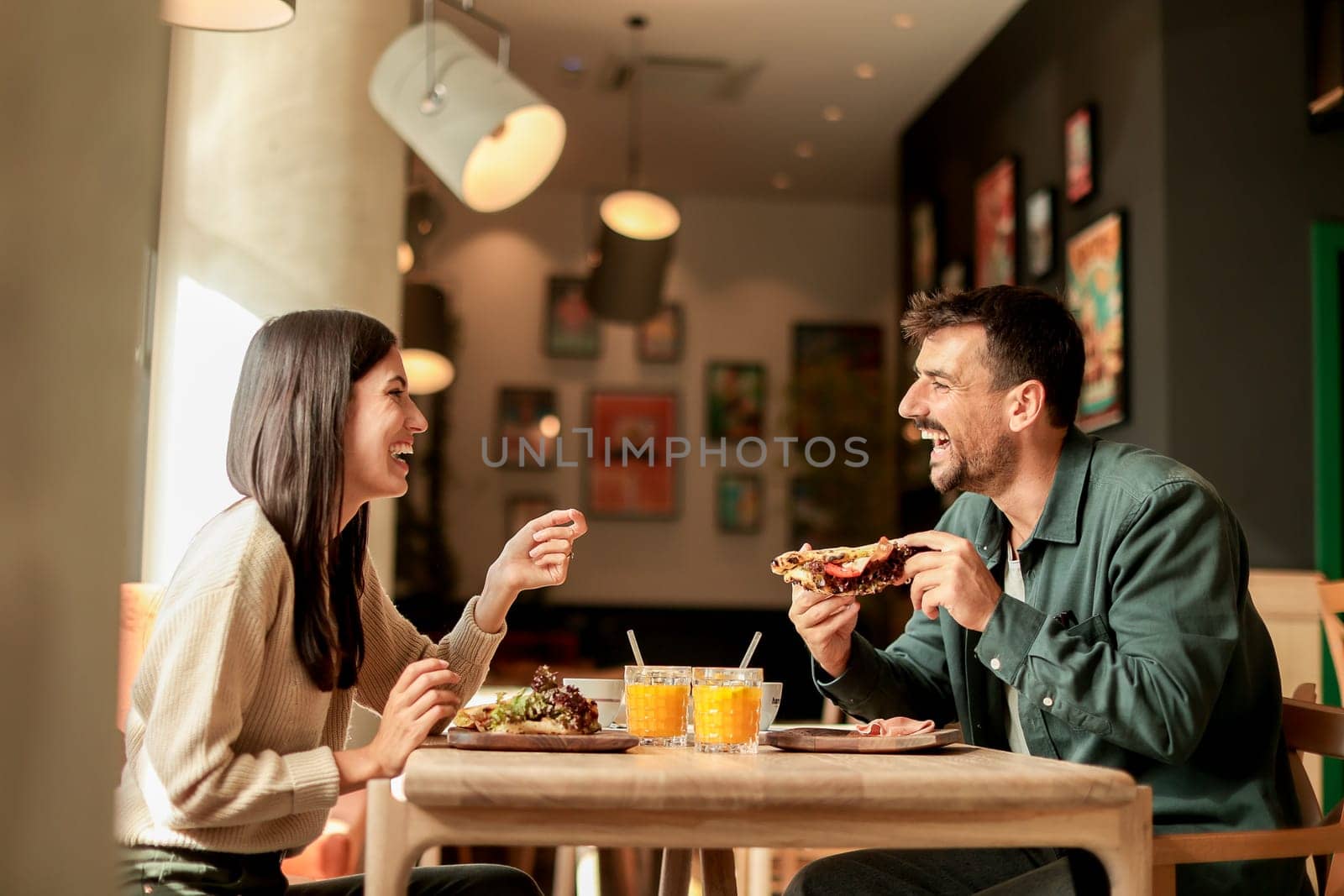 Couple shares laughter and delicious food while sitting at a wooden table in a bright cafe, surrounded by greenery and other patrons enjoying their meals