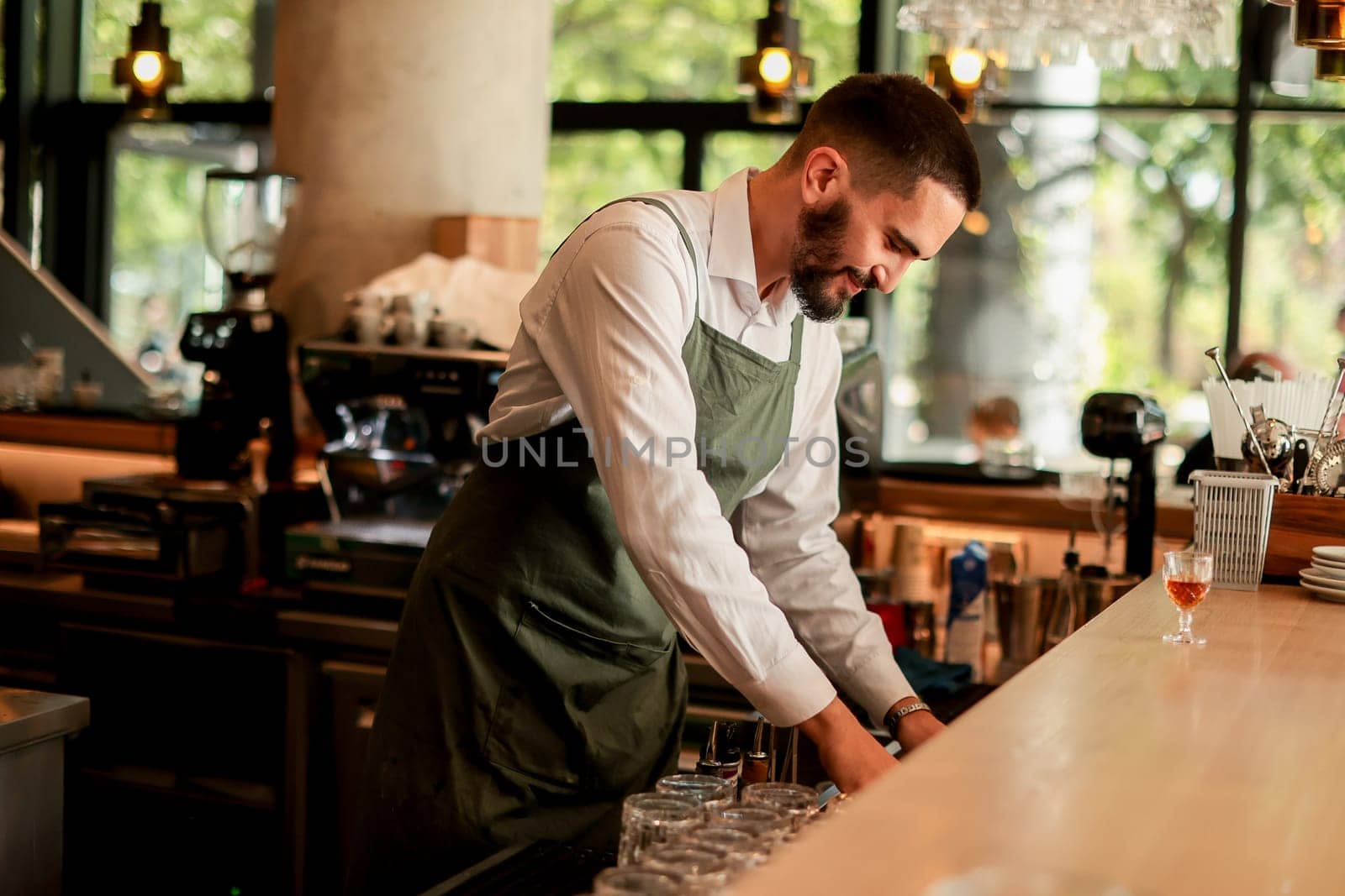 With a warm smile, the bartender skillfully crafts cocktails in a lively bar, where sunlight floods through large windows, creating a vibrant atmosphere