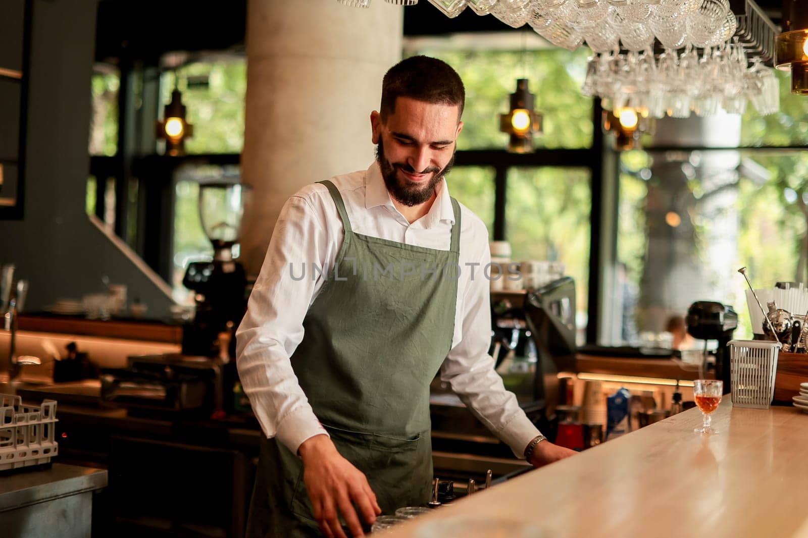 With a warm smile, the bartender skillfully crafts cocktails in a lively bar, where sunlight floods through large windows, creating a vibrant atmosphere
