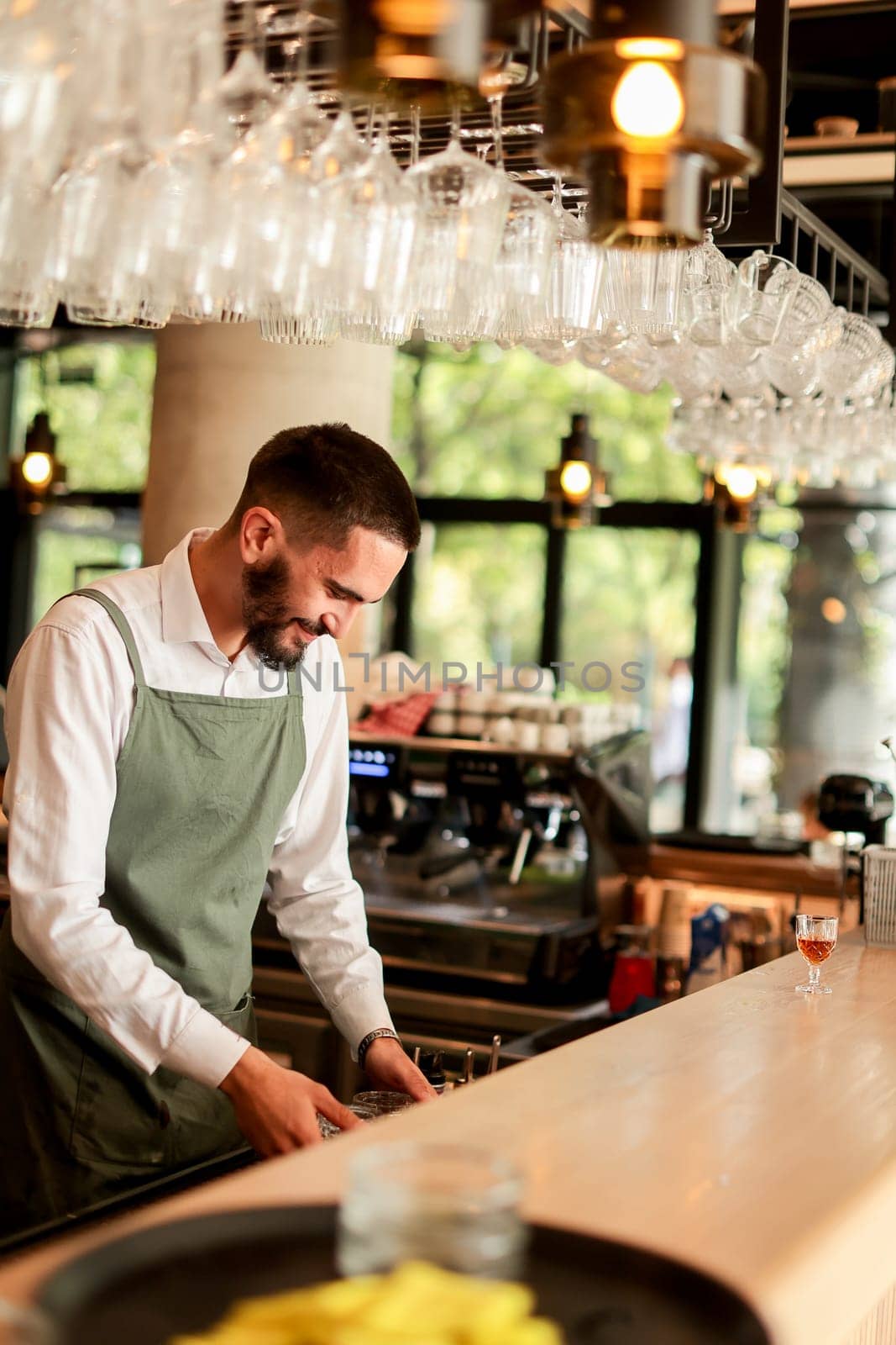 With a warm smile, the bartender skillfully crafts cocktails in a lively bar, where sunlight floods through large windows, creating a vibrant atmosphere