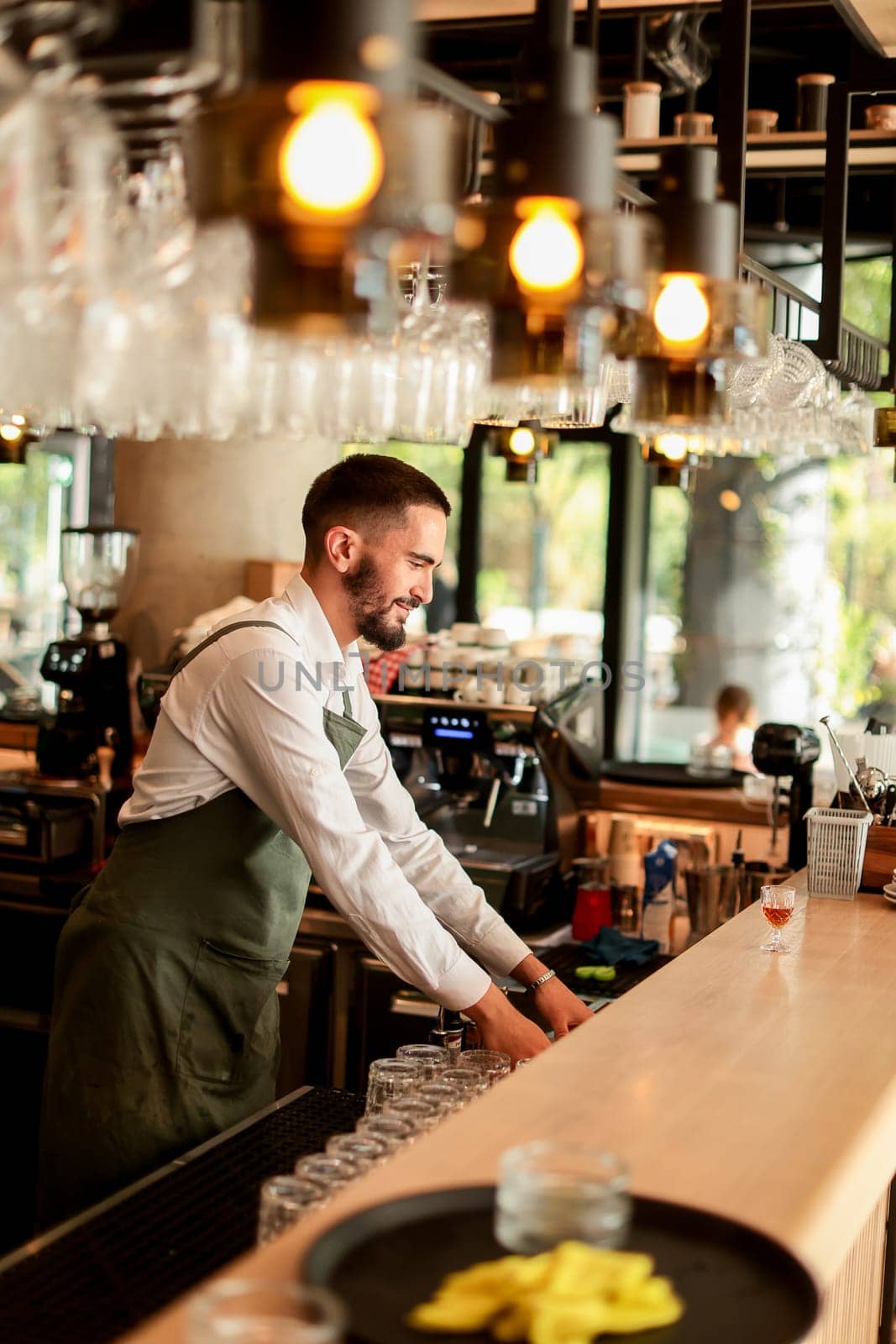 With a warm smile, the bartender skillfully crafts cocktails in a lively bar, where sunlight floods through large windows, creating a vibrant atmosphere