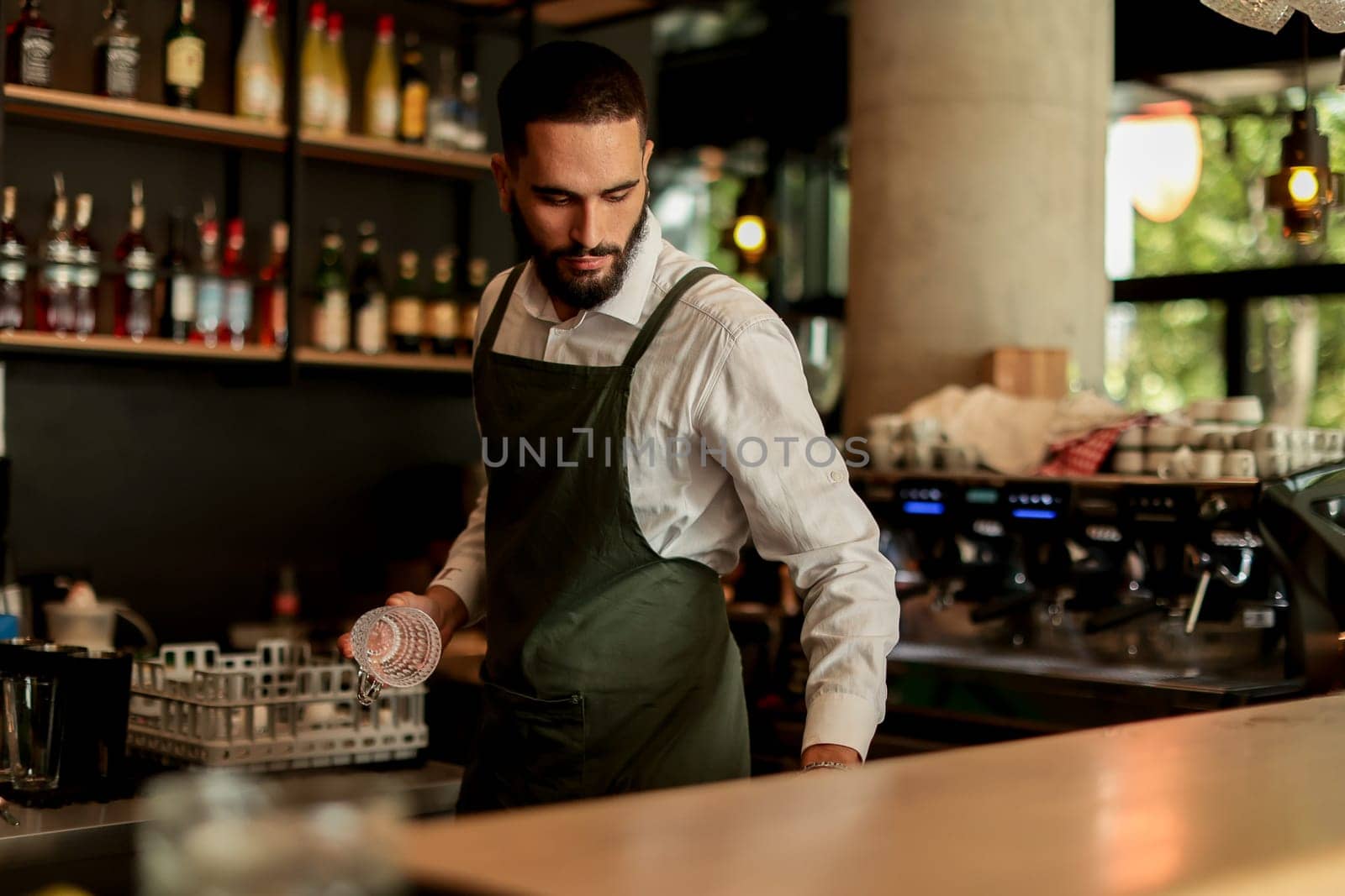 Bartender in an elegant apron meticulously arranges glassware while serving patrons in a lively café environment, highlighting craft and hospitality