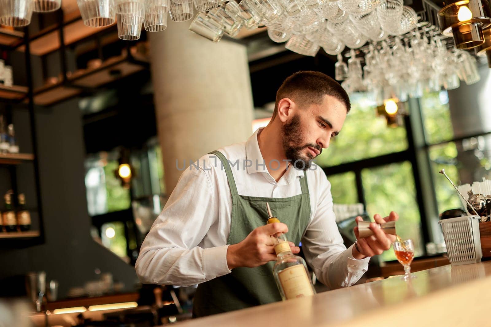 Bartender concentrates as he skillfully crafts a cocktail, surrounded by an array of glassware in a modern bar filled with natural light, creating an inviting atmosphere