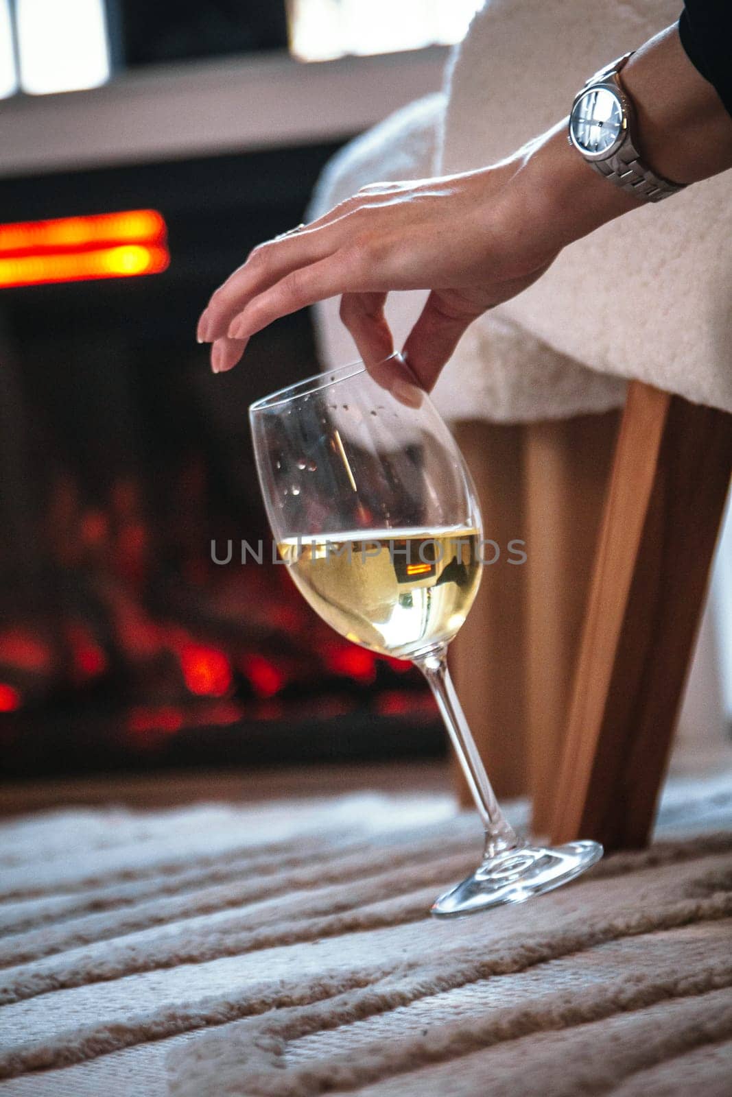 girl with watch on hand holds glass of wine with two fingers, fireplace burning in background in modern interior, romantic atmosphere of relaxation. Warm atmosphere