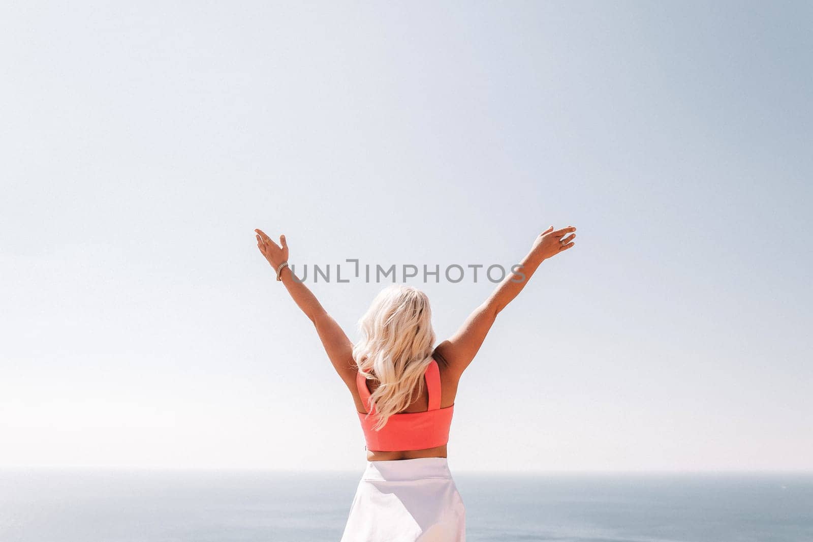 A woman in a pink top and white skirt is standing on a cliff overlooking the ocean. She is smiling and she is happy