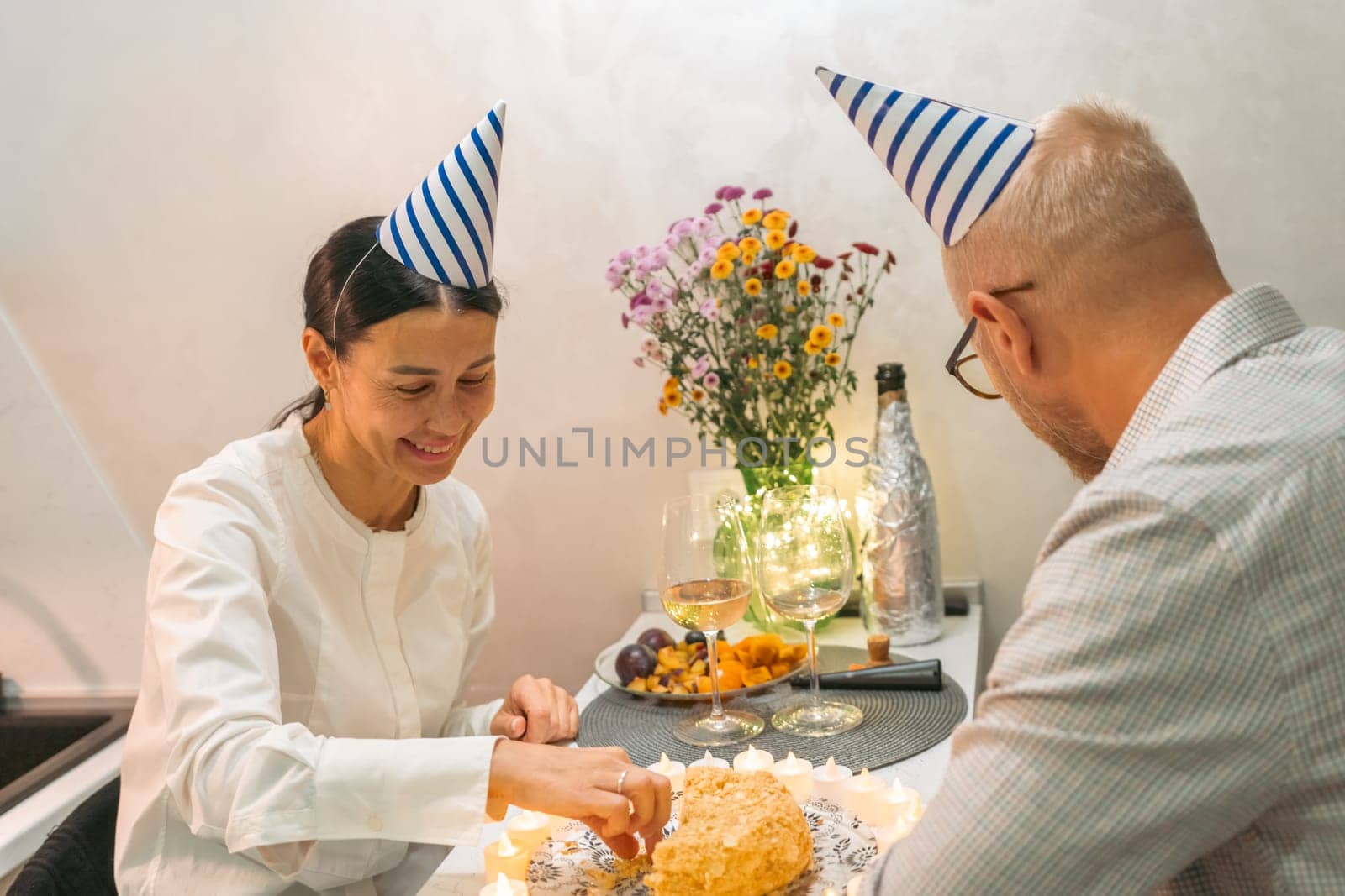 Here's a caption under 150 characters describing the image: A man and woman, wearing party hats, celebrate with food and drinks at an intimate table setting.