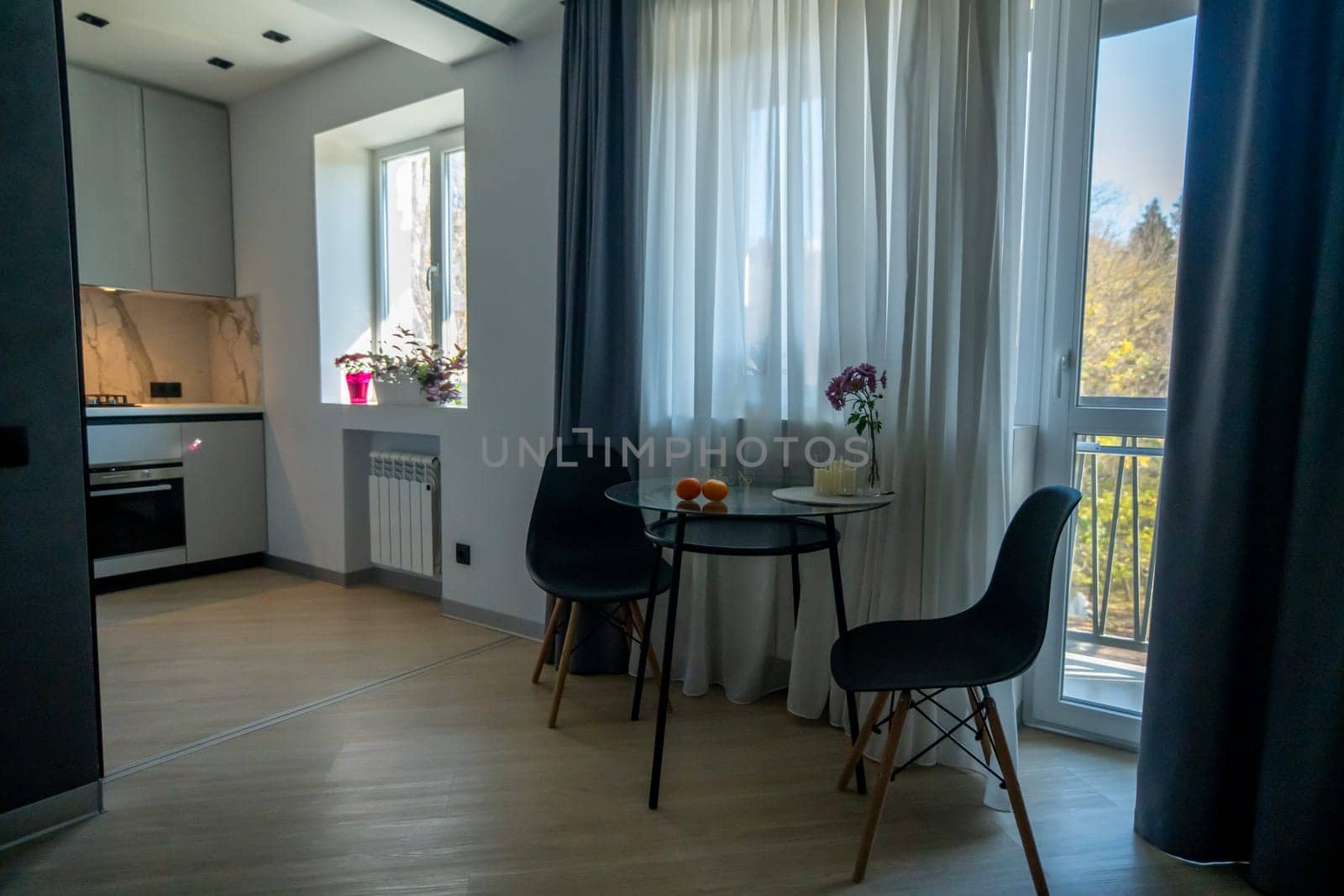 Modern apartment interior: kitchen, dining area with small table and two black chairs near window with sheer curtains, overlooking a balcony and trees.