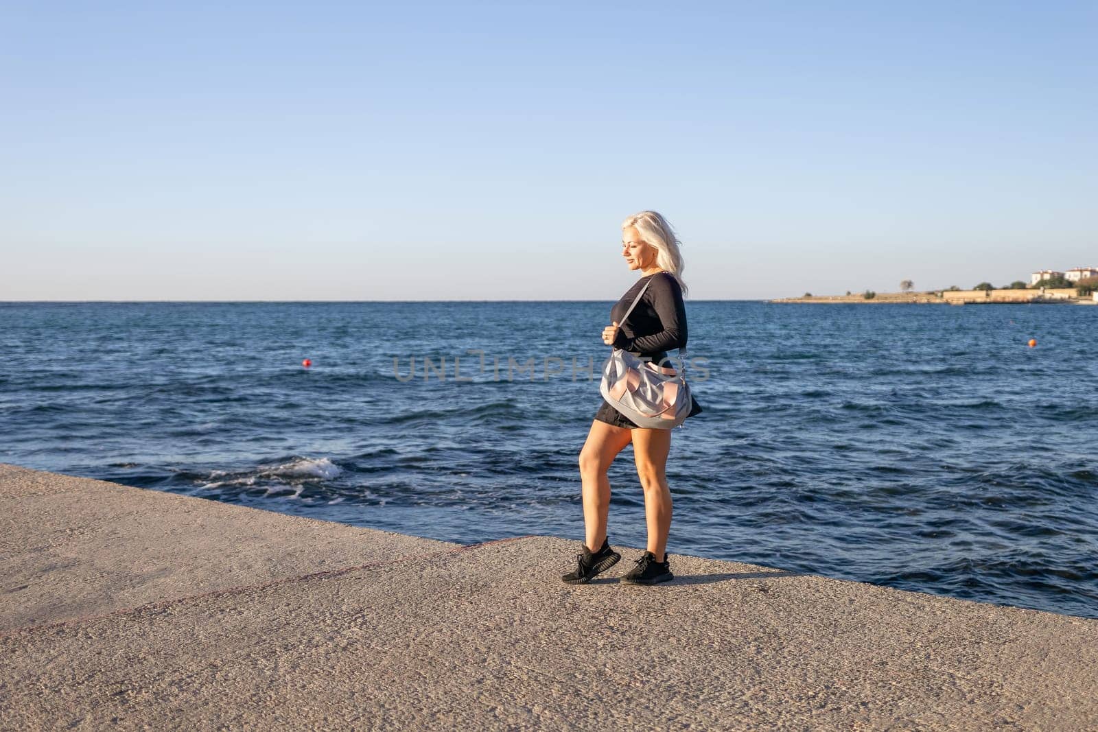 A blonde woman in a black shirt and skirt stands on a concrete pier, looking out at the blue ocean.