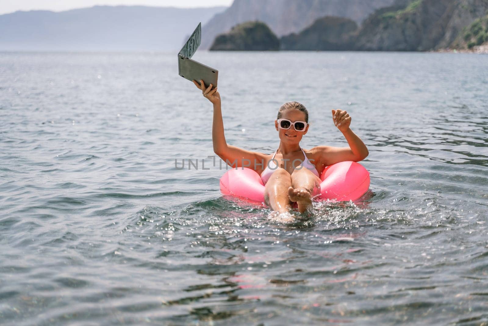A woman is floating in a pink inflatable raft with a laptop in her hand. She is smiling and she is enjoying her time in the water