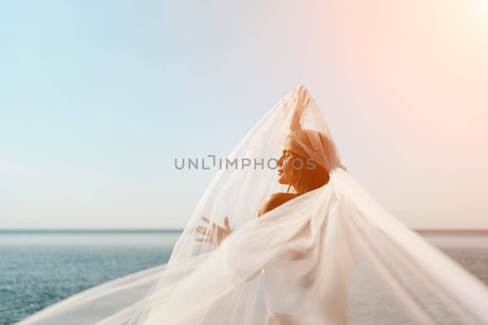 A woman stands on a beach with a white veil over her head. The scene is serene and peaceful, with the woman's gaze directed towards the water
