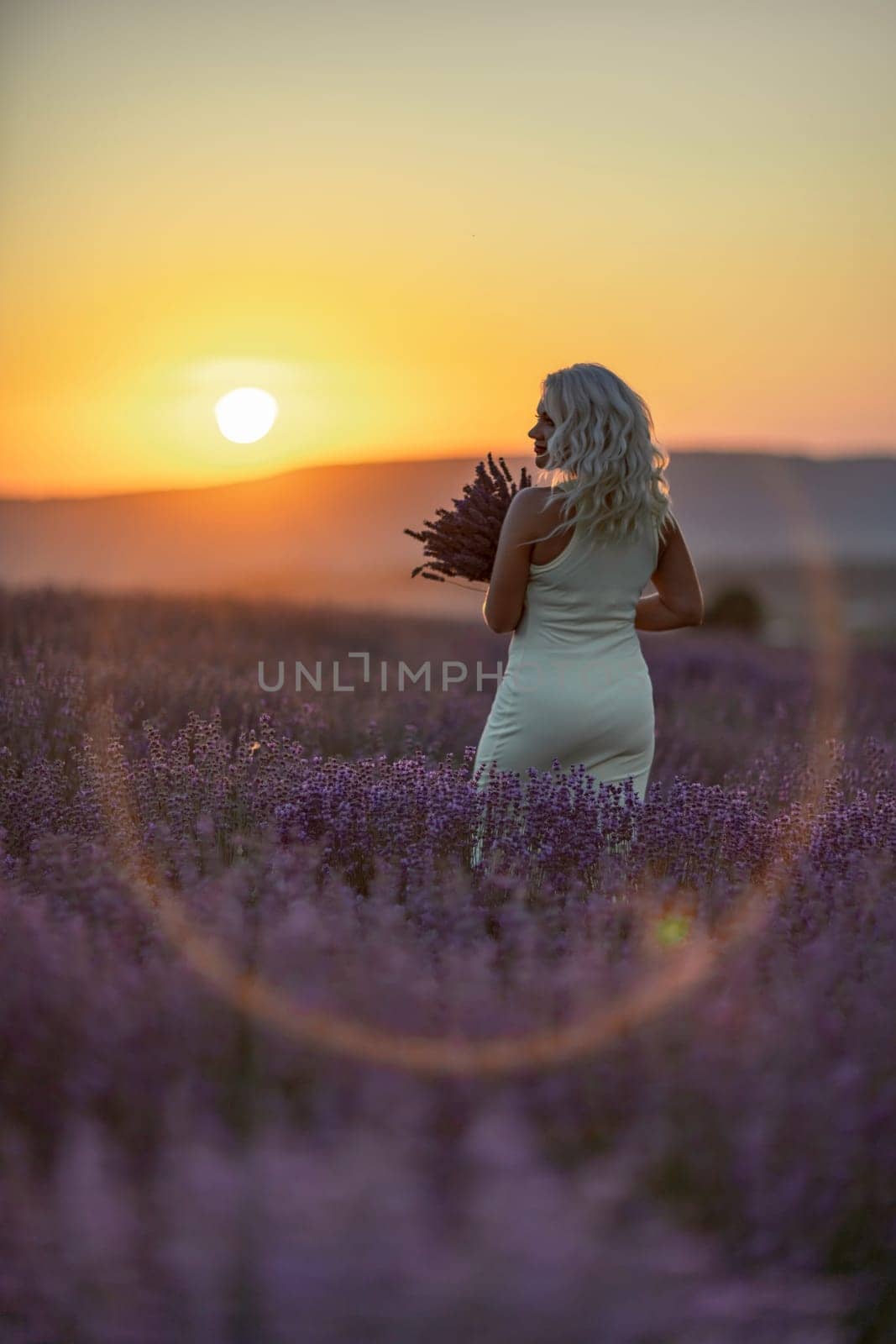 Blonde woman poses in lavender field at sunset. Happy woman in white dress holds lavender bouquet. Aromatherapy concept, lavender oil, photo session in lavender.