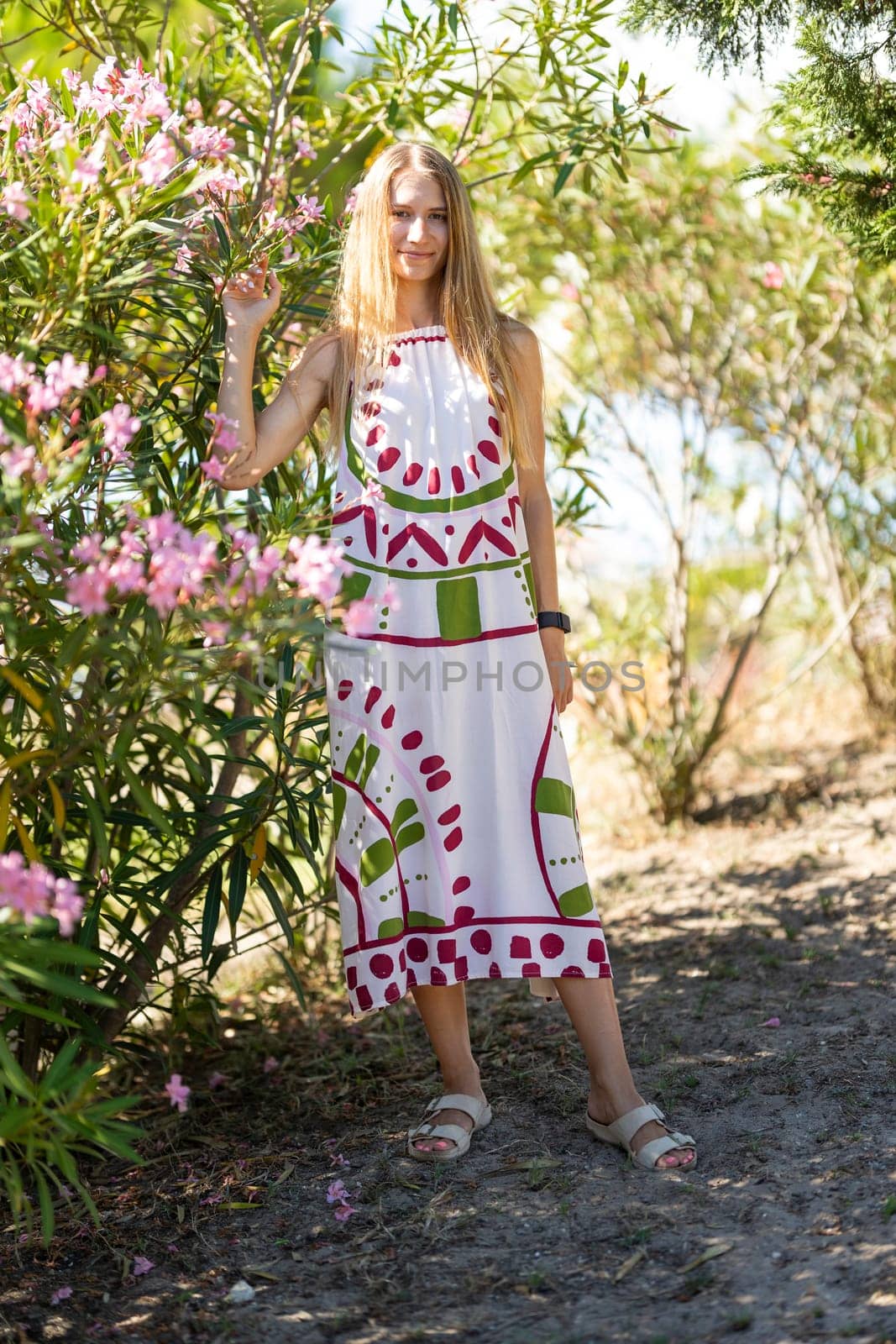 Young blonde woman is posing in a botanical garden, wearing a long colorful dress and touching oleander flowers