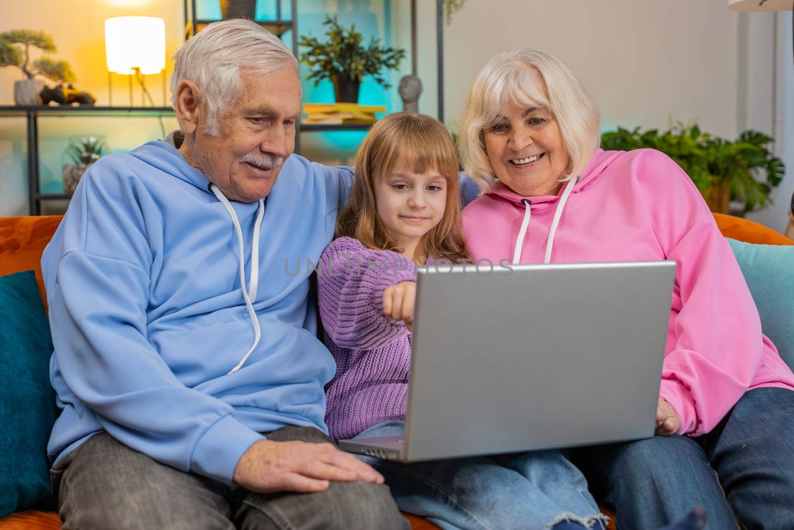 Senior Caucasian couple man and woman with child girl granddaughter spending time at home together, sitting on sofa in the living room, using laptop computer. Watching videos playing games on weekend.