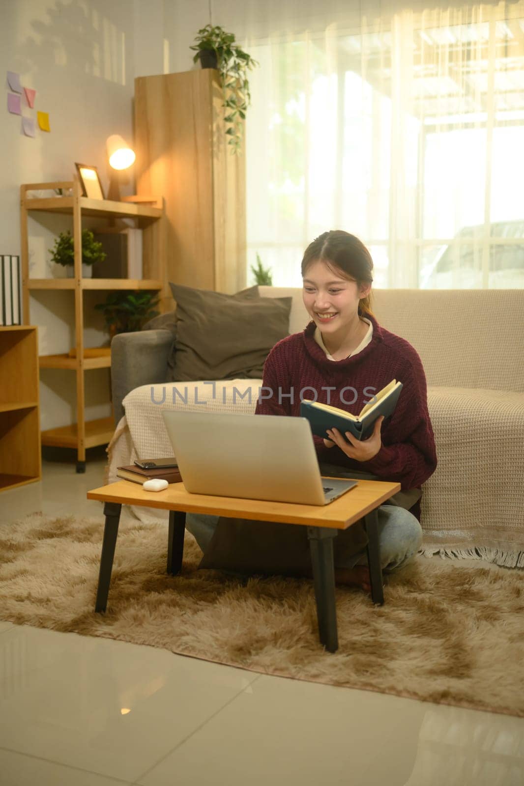 Young female student holding notebook and using laptop in living room.