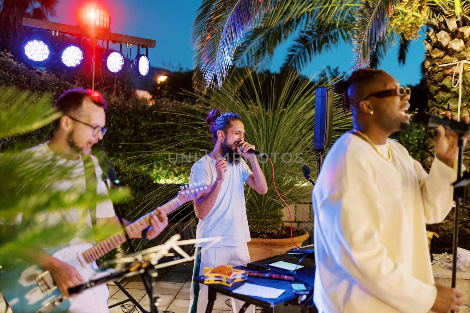 Male musical group sings into microphones, standing on the restaurant site among green trees. High quality photo