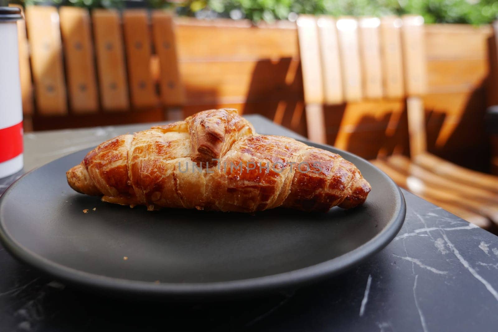 A flaky croissant is served on a black plate under warm sunlight.