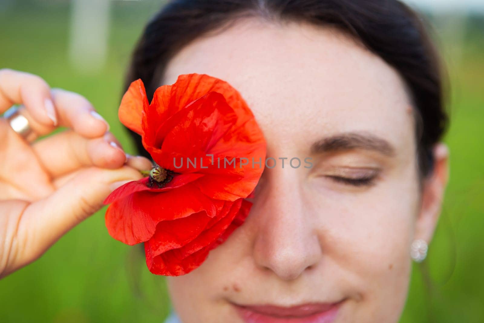 A young woman gently holds a vibrant red poppy flower to her closed eye, creating a serene and beautiful moment outdoors.