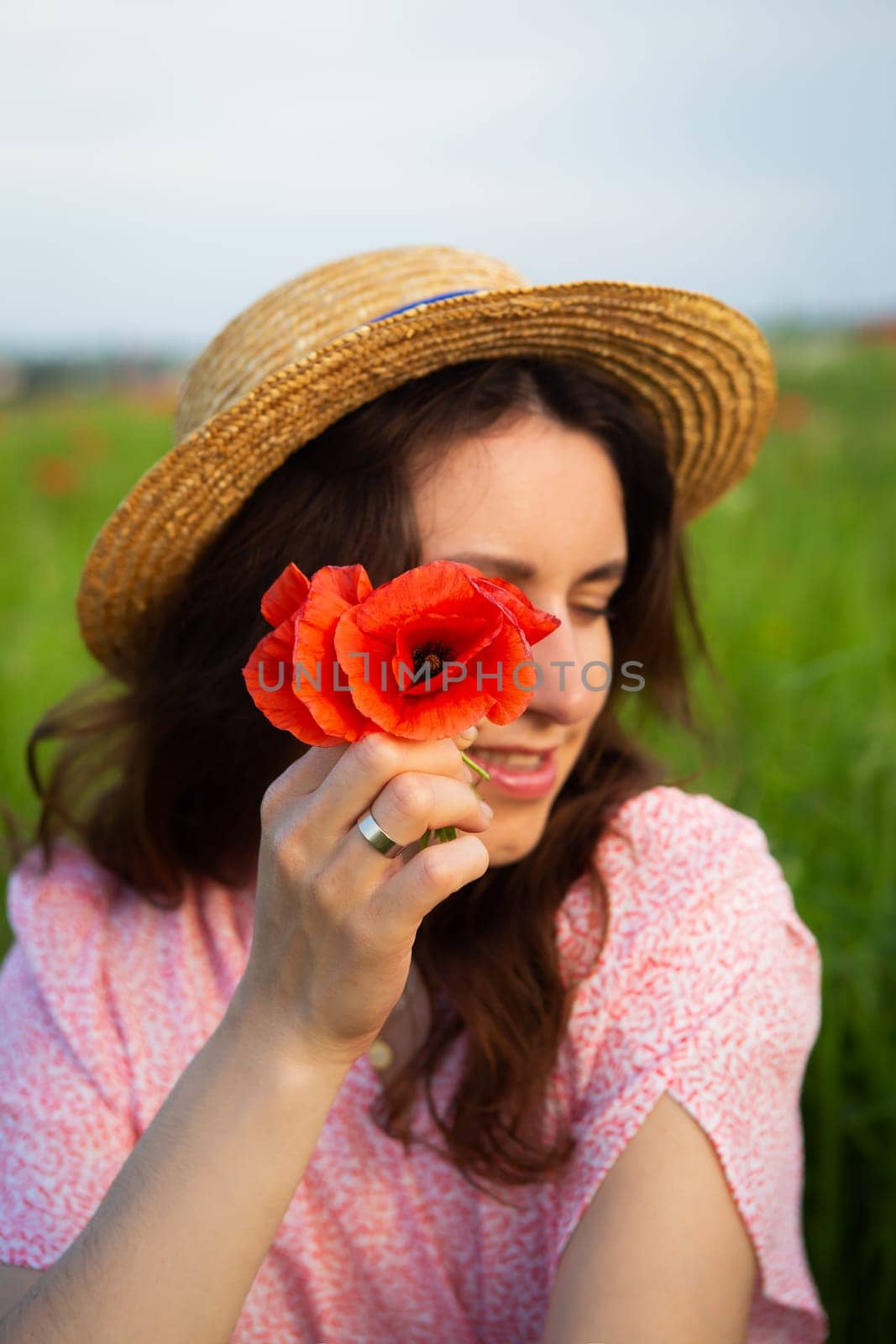 A young woman in a straw hat holds a vibrant red poppy flower near her face in a sunny field.