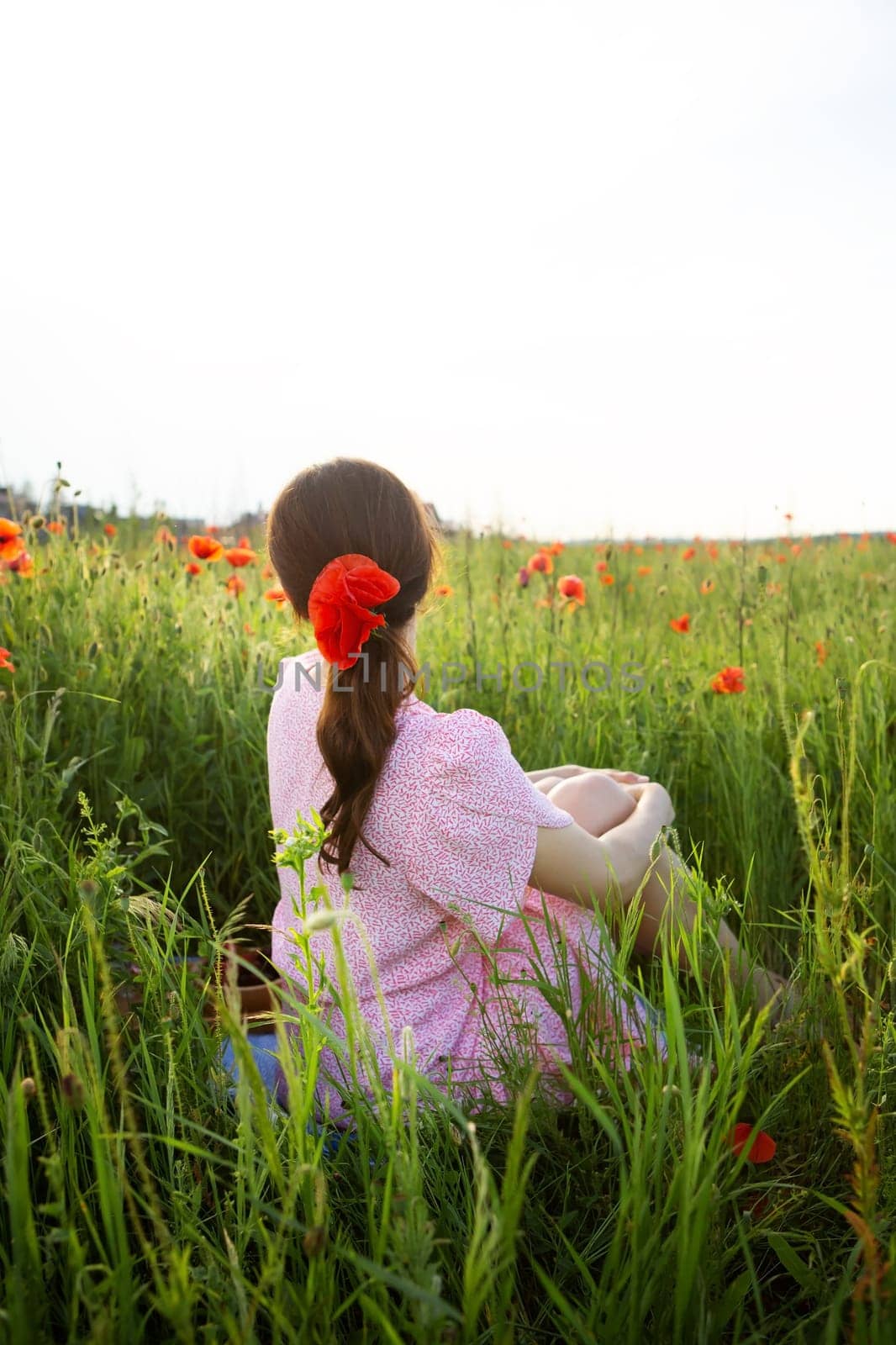 A young woman with a poppy in her hair sits peacefully in a vibrant poppy field, enjoying the summer sun.