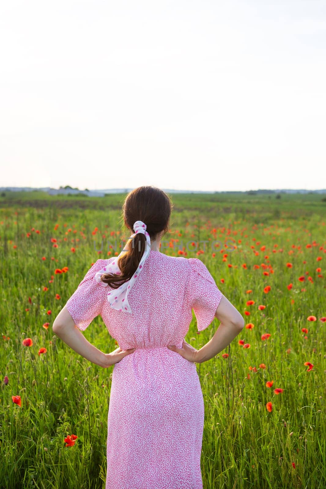 A woman in a pink floral dress stands in a poppy field, gazing at the horizon on a sunny day.