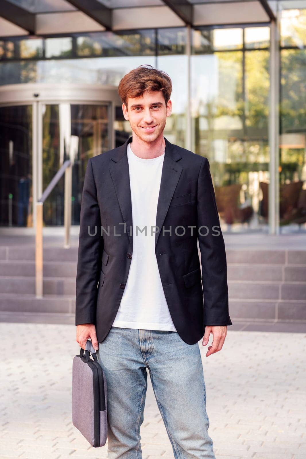 young adult man in casual suit standing looking at camera holding a briefcase in front of an office building, concept of entrepreneur and business