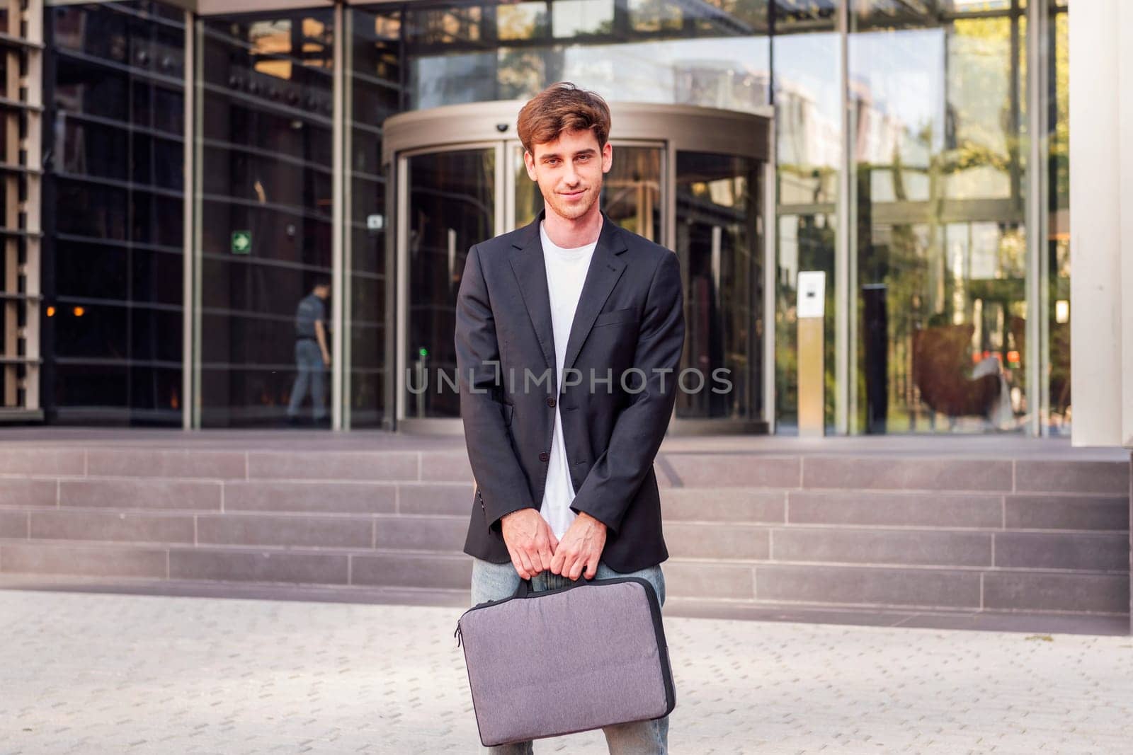 young adult man in casual suit standing looking at camera holding a briefcase in front of an office building, concept of entrepreneur and business, copyspace for text