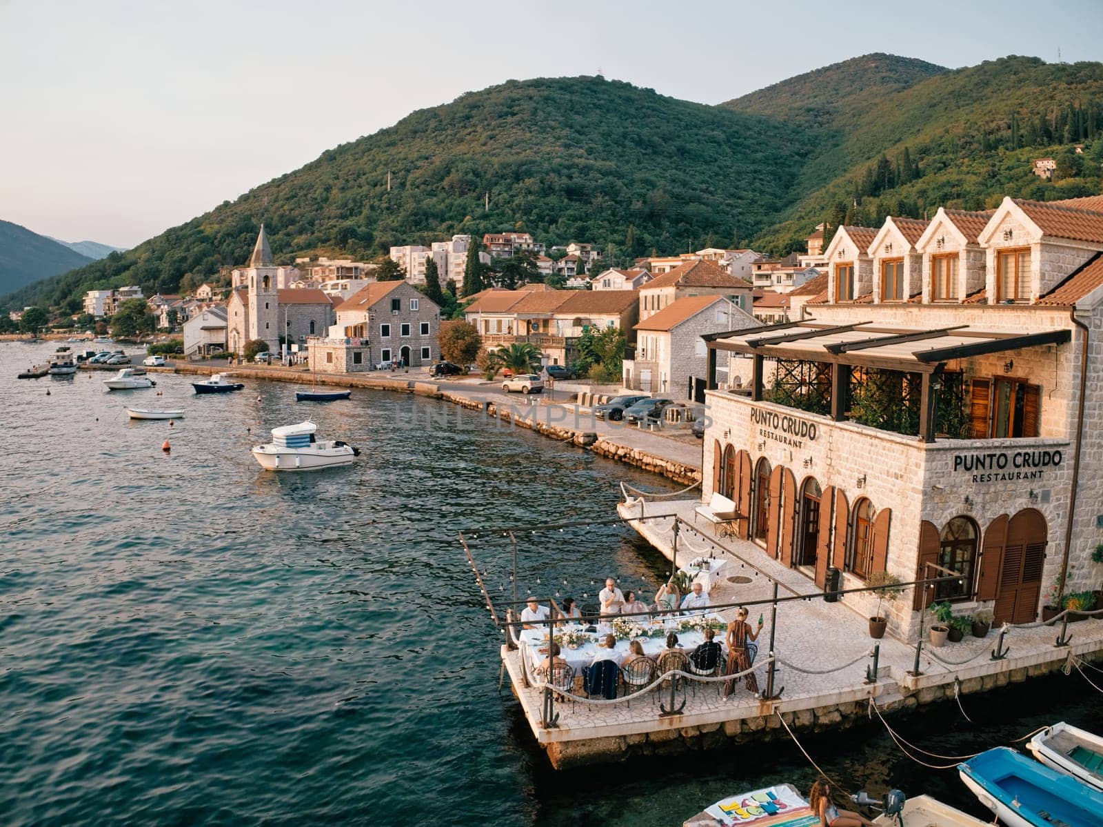Guests are sitting at a festive table on the terrace by the sea of the Punto Crudo restaurant. Tivat, Montenegro. High quality photo