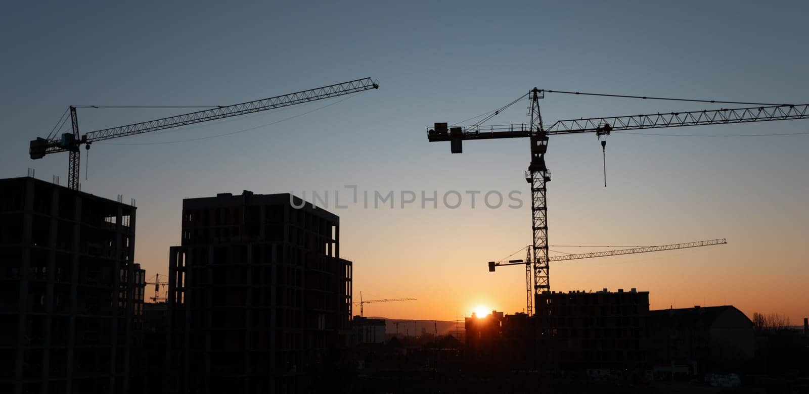 Construction cranes tower over a developing urban area as the sun sets, casting a golden glow on the buildings. The cranes are actively working, signifying ongoing projects.