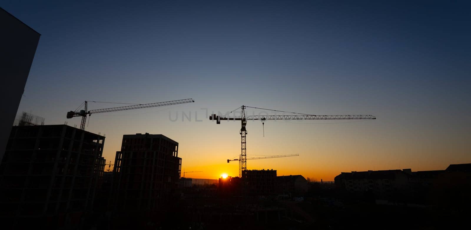 A vivid sunset casts warm hues over a construction site, where cranes stand tall against the skyline. The silhouettes of buildings create a dramatic contrast in the fading light.