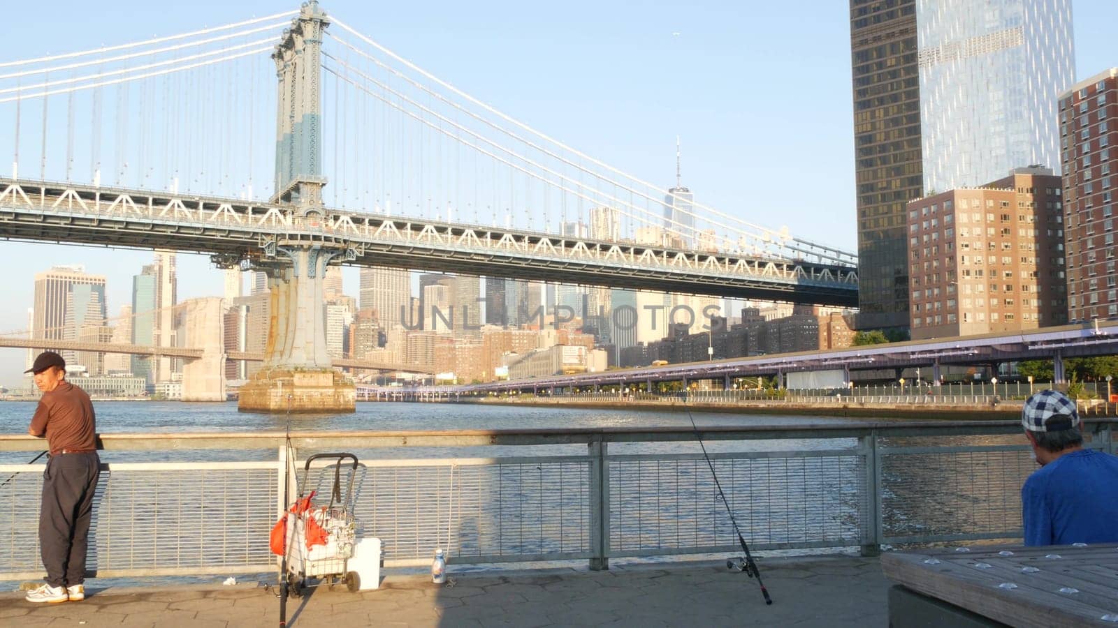New York City, United States - 7 Sept 2023: Manhattan Bridge. Fisherman near Pier 35 swings, waterfront downtown skyline cityscape. World Trade Center skyscraper, people fishing and morning sunset.