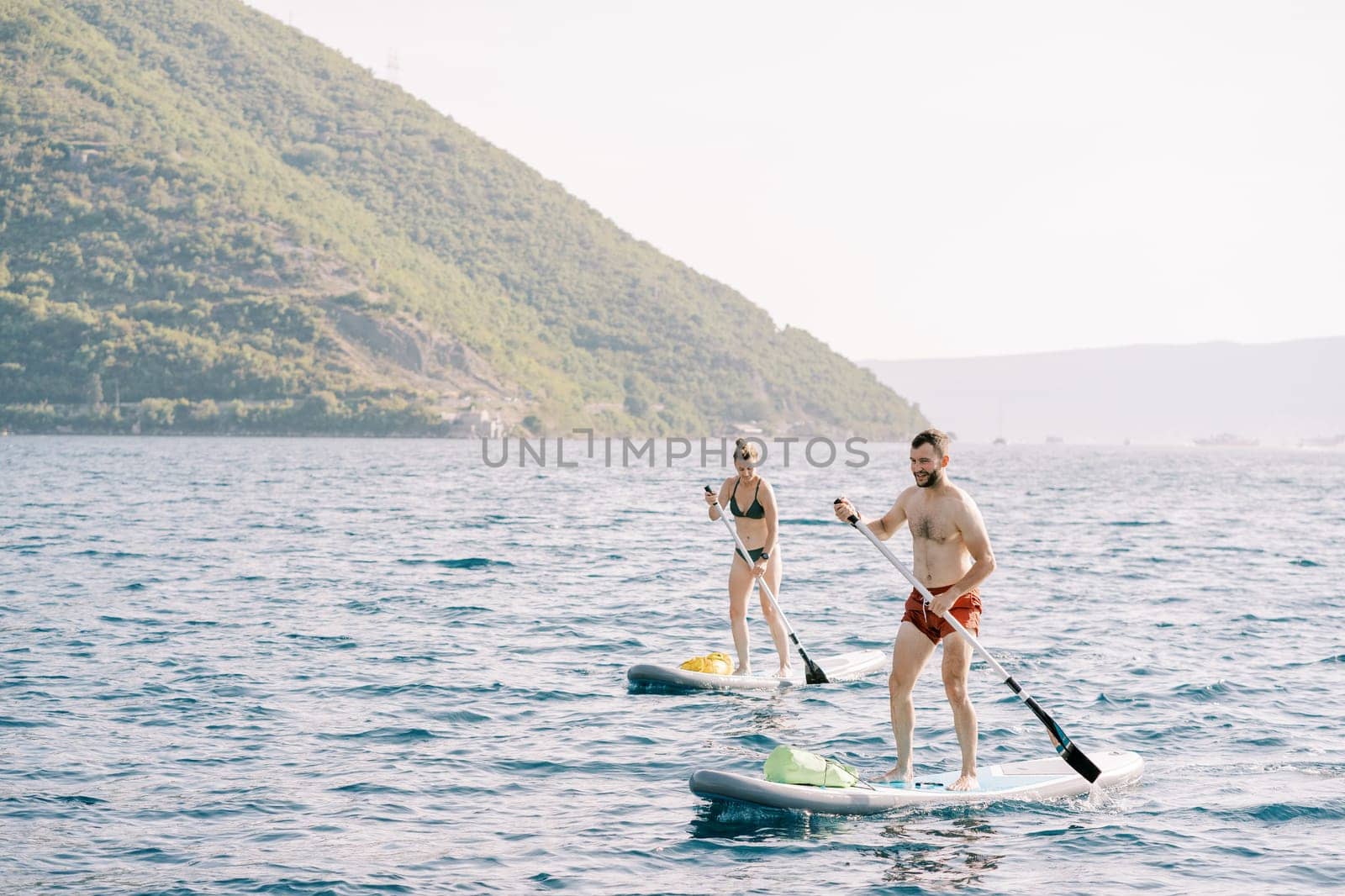 Man and woman swim standing on boards rowing oars in the sea against the backdrop of mountains. High quality photo