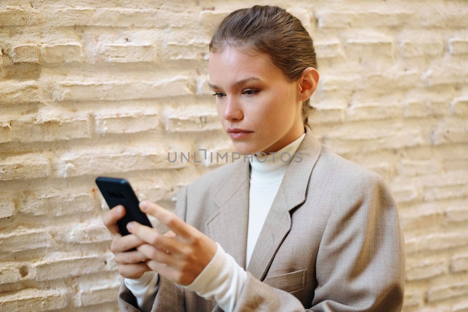 A young woman in a stylish blazer stands confidently against a textured wall, focusing on her smartphone, embodying the essence of modern lifestyle and technology in todays fastpaced world