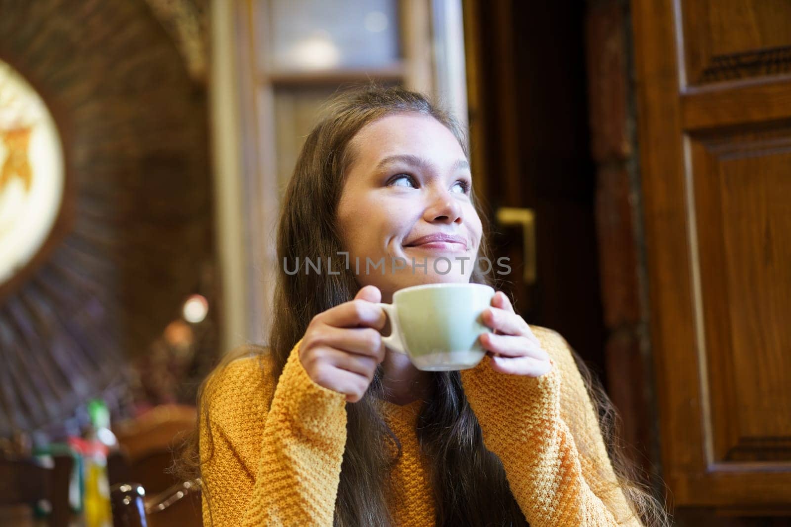 A cheerful young woman with long hair is happily enjoying a warm cup of coffee in a cozy, inviting cafe, radiating an aura of warmth and pure happiness that brightens the atmosphere around her