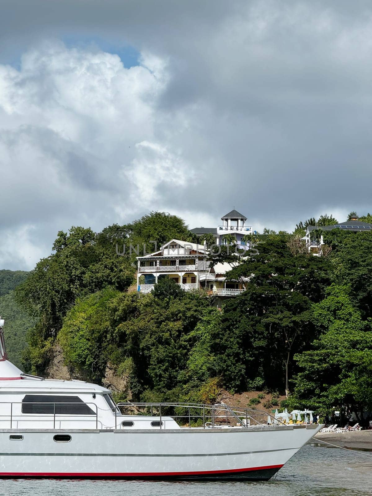 White yacht anchored near a lush tropical coastline with hillside villas and dramatic clouds overhead, evoking a serene vacation vibe.