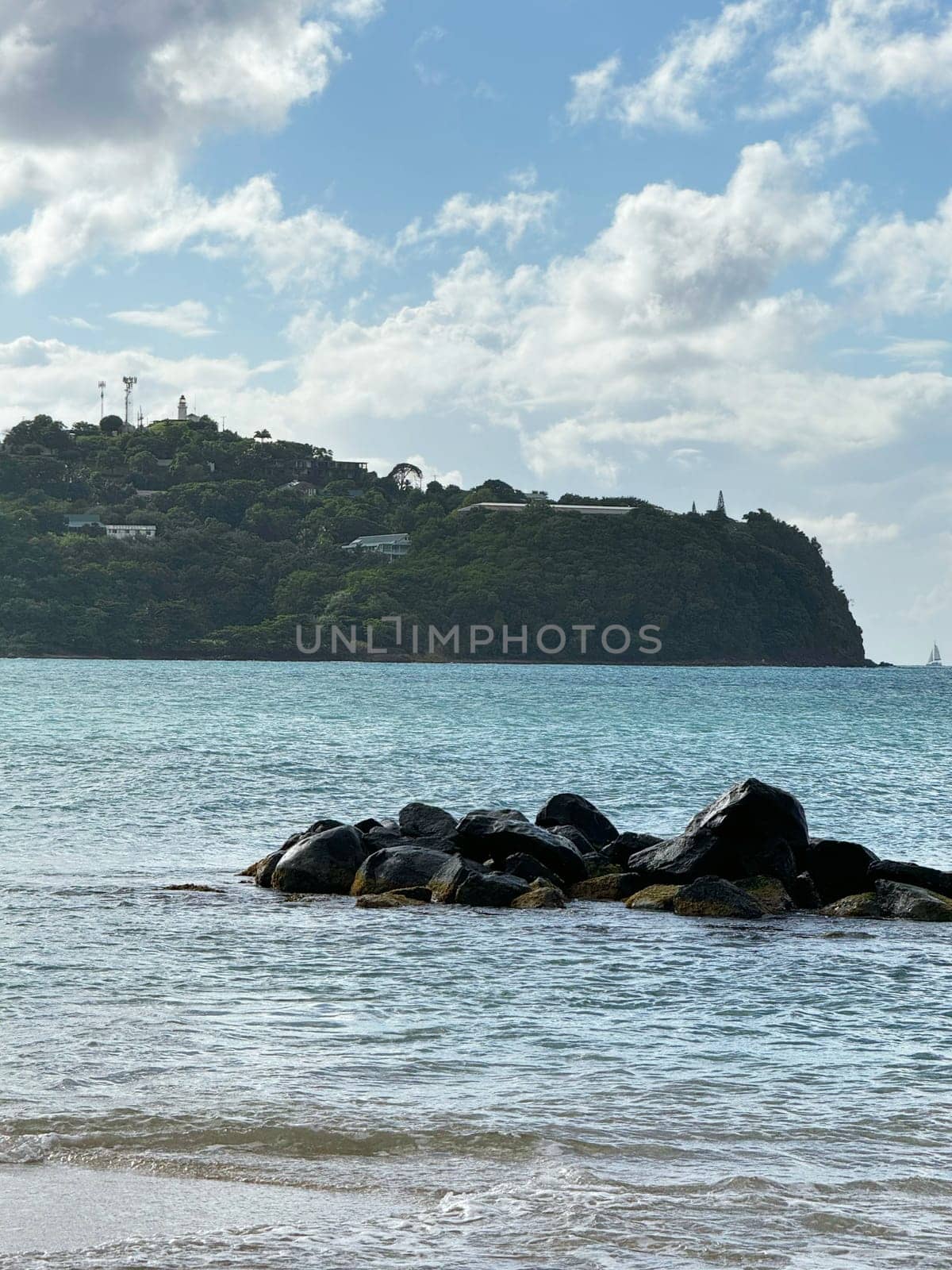 Peaceful coastal scene with dark rocks in the shallow water, clear turquoise sea, and a lush green hillside under a partly cloudy sky in the tropical Caribbean.