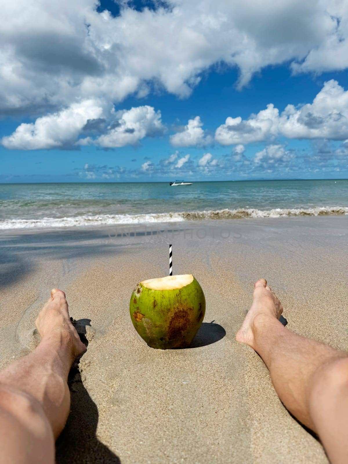 Person relaxing on a sandy tropical beach with a fresh coconut drink placed between their legs, facing the calm sea and vibrant sky filled with fluffy white clouds.