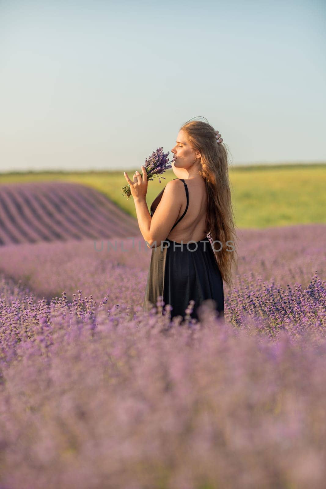 woman stands in a lavender field of purple flowers, holding a bouquet of flowers. The scene is serene and peaceful, with the woman taking a moment to enjoy the beauty of nature