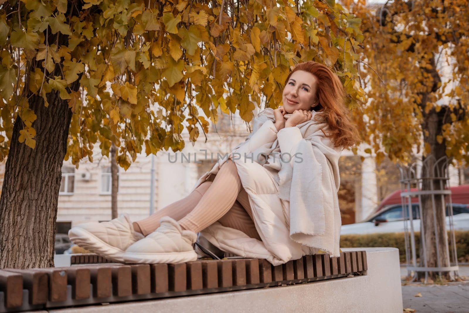Redhead woman in a light coat sits on a park bench under autumn leaves, smiling. She wears sneakers.