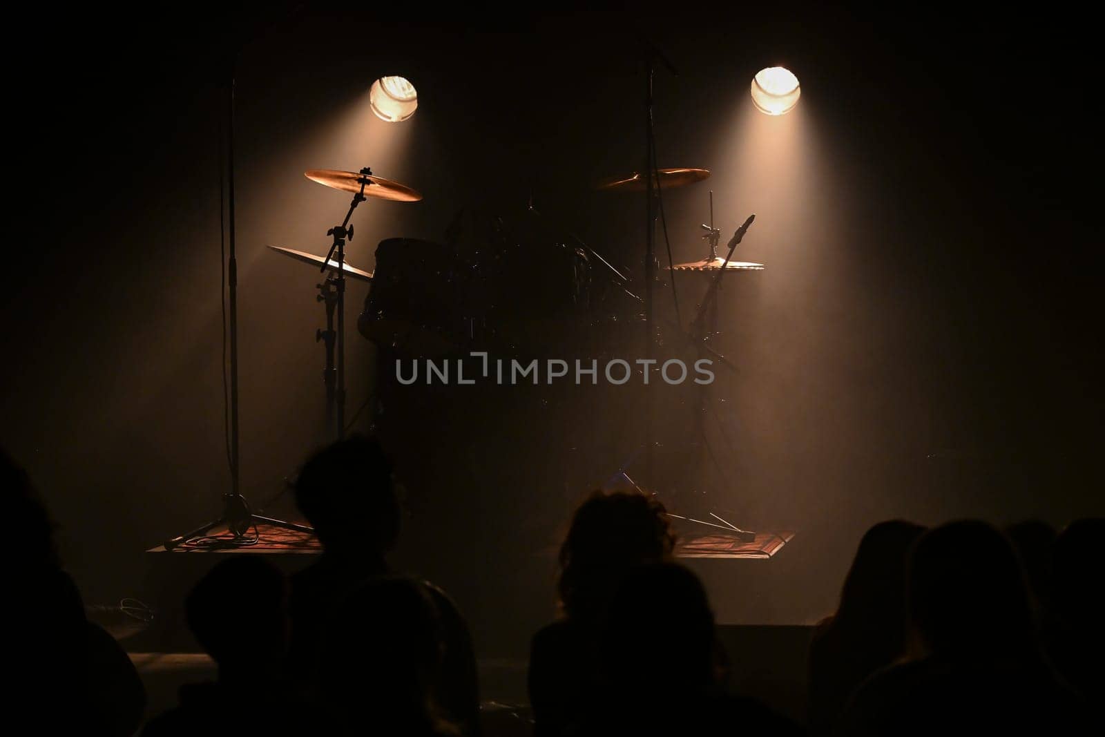 Bright stage lights illuminate the drum set as an excited crowd waits for the performance to begin, creating an atmosphere full of energy and anticipation.