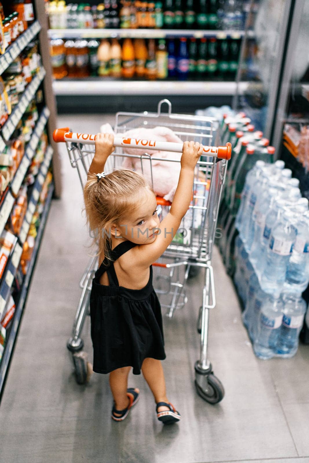 Little girl pushes a grocery cart around the store and looks back. Back view. High quality photo