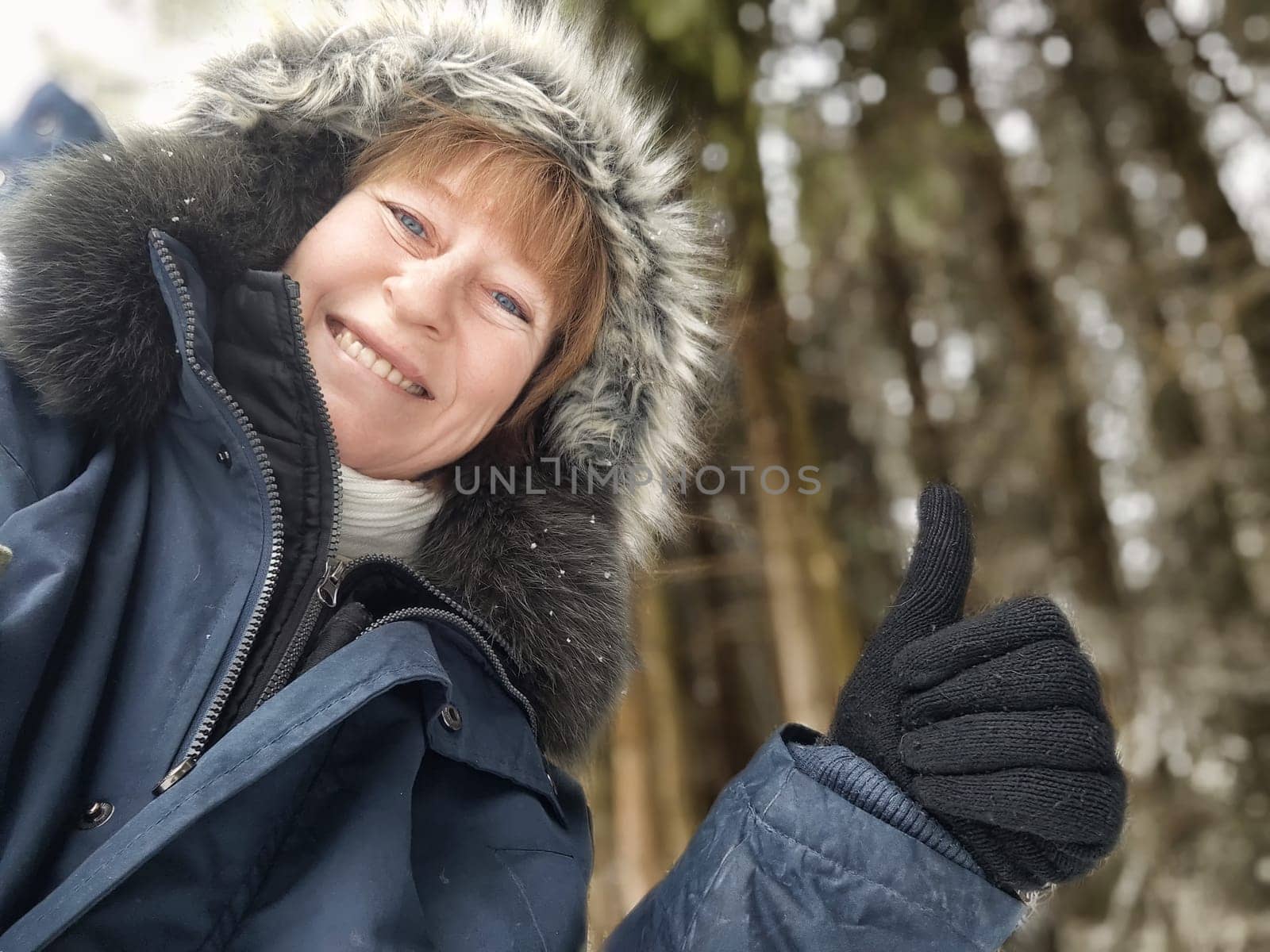 A middle-aged tourist happily poses for a selfie, giving a thumbs up while dressed warmly in a snowy forest, capturing her travel joy.