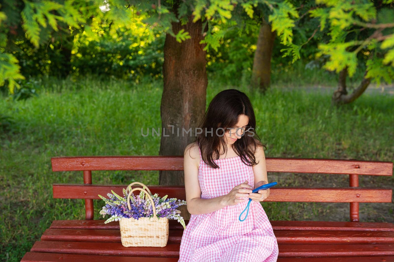 A young woman in a pink gingham dress sits on a red park bench, using her phone, with a basket of lupines beside her.