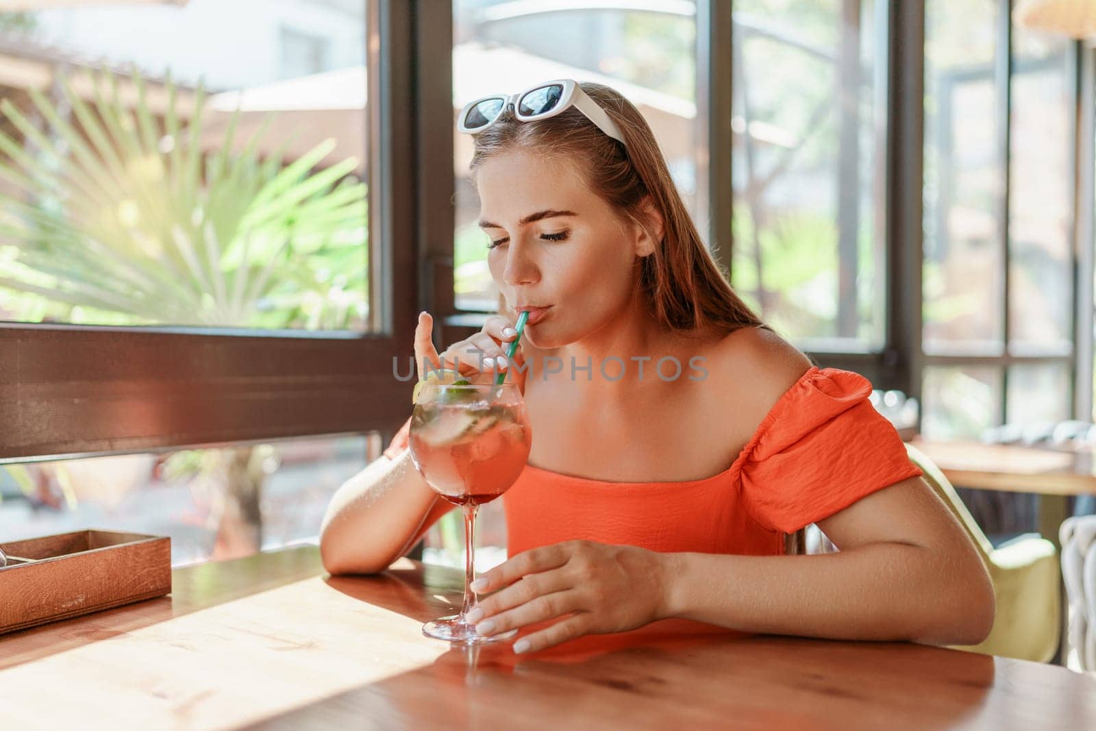 woman in a cafe seated, holding beverage. Dressed in an orange dress with her hair down. Bright interior, large windows allow natural light. Beverage consumption for refreshment