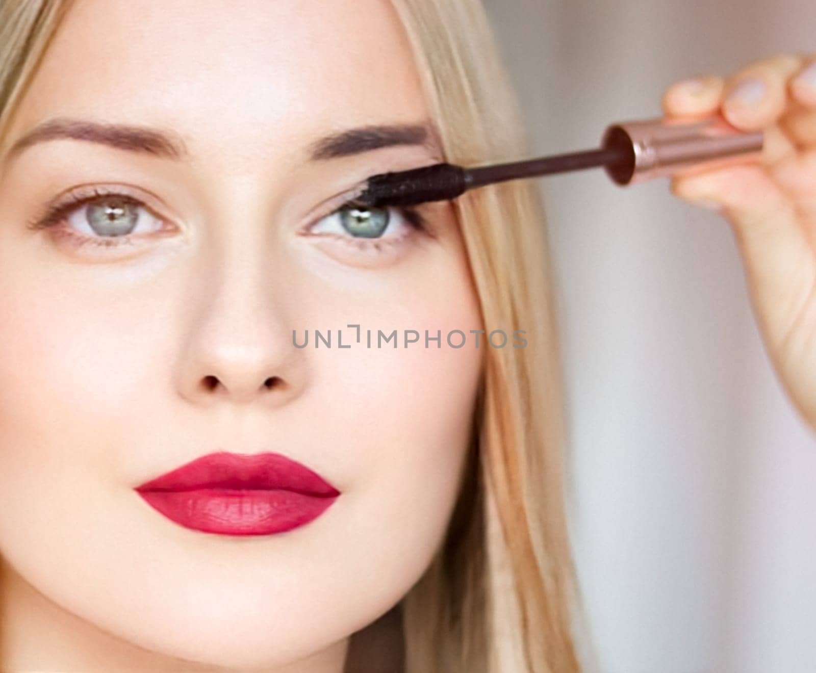 Woman applying mascara with confidence, defined brows and red lipstick. Blonde beauty focused on her eye makeup, cosmetics look